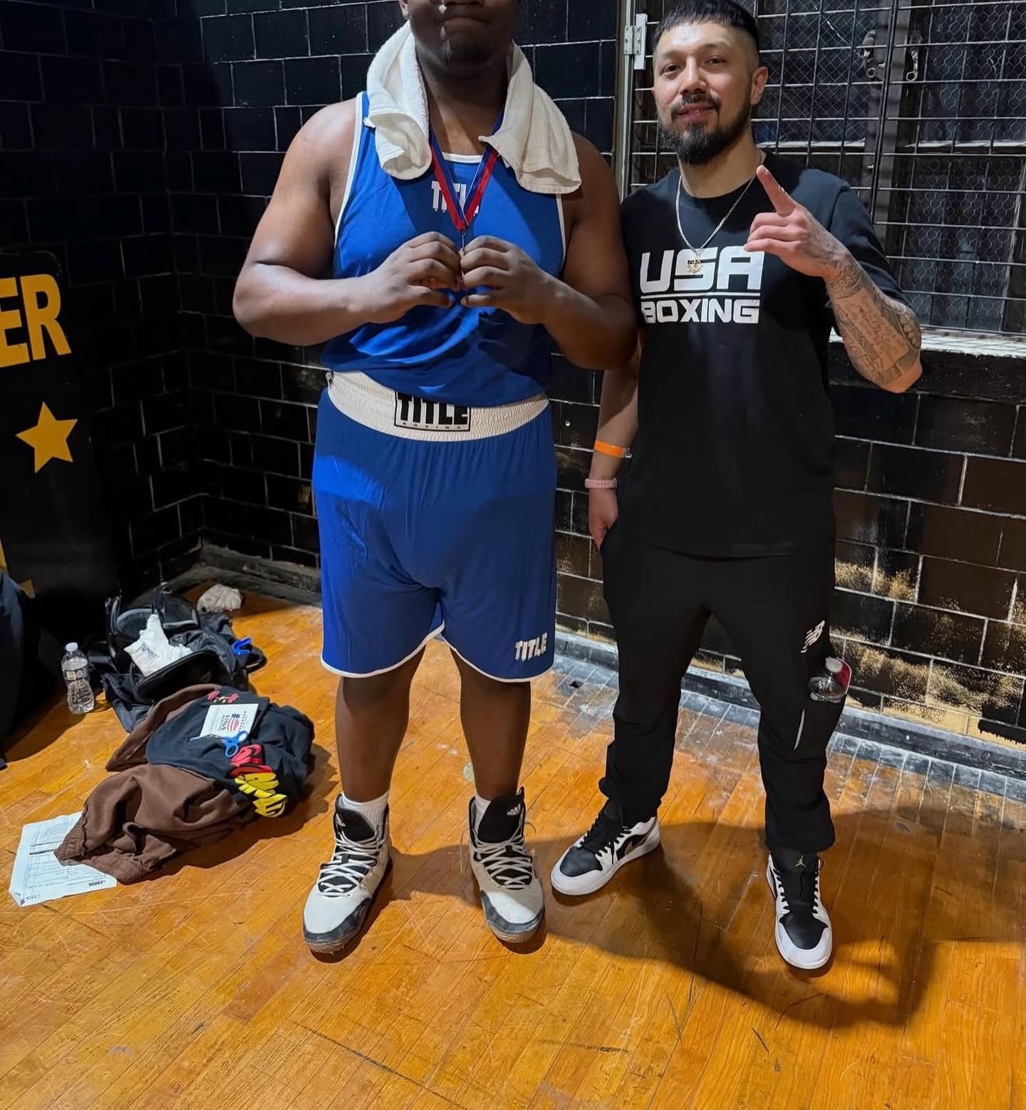 A boxer in a blue TITLE uniform standing with a coach in a USA Boxing tee backstage.