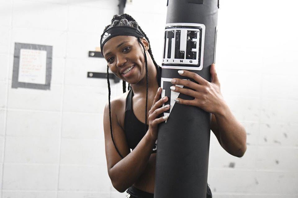 An adult woman boxer in a sports bra and headband smiling beside a black TITLE heavy bag in the gym.