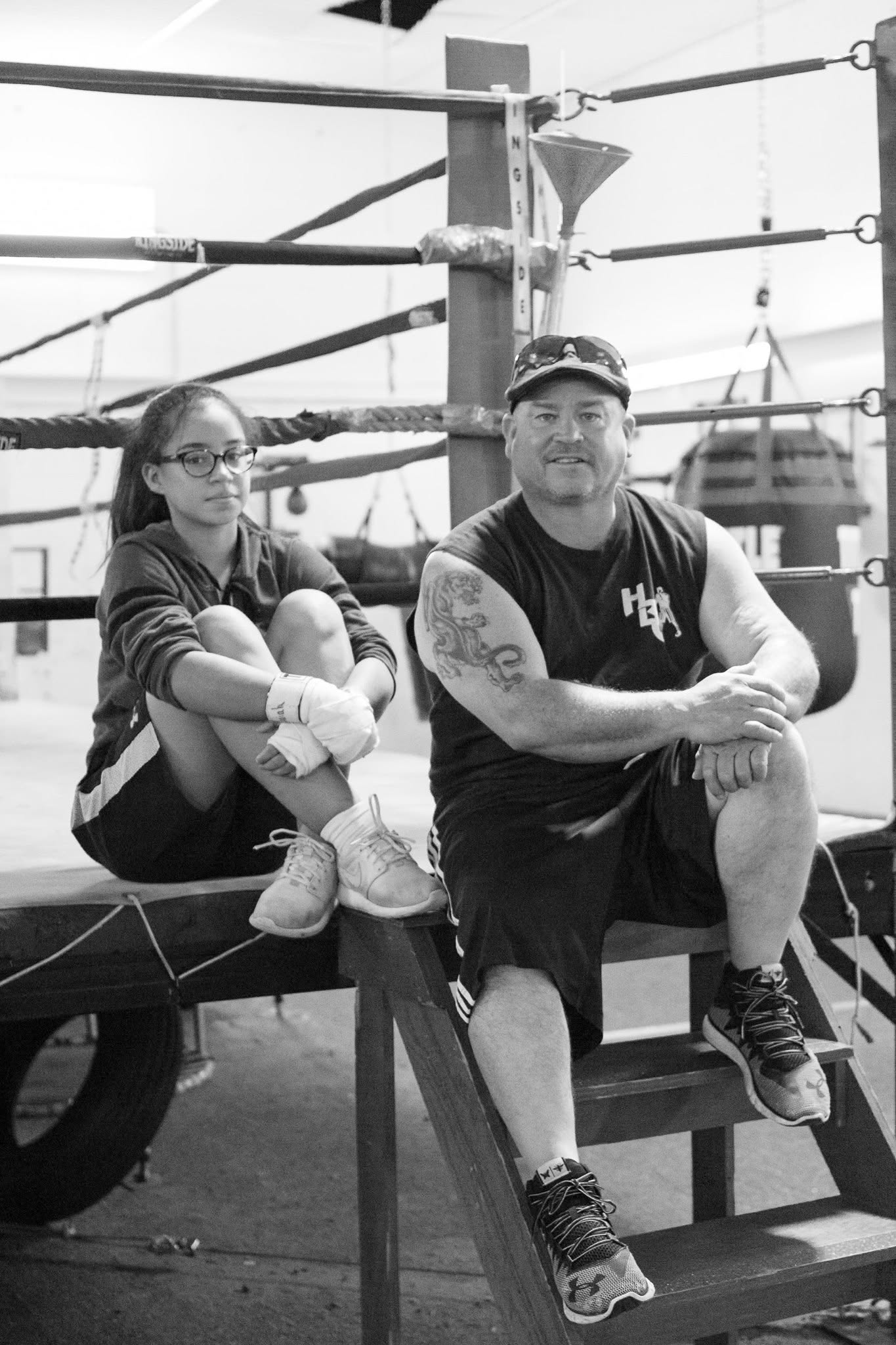 Black-and-white photo of a coach in an HBC tank sitting on the ring steps beside a young female boxer with wrapped hands.