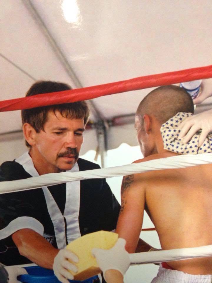 A cornerman in a black robe tending to a fighter between rounds at an outdoor tented ring.