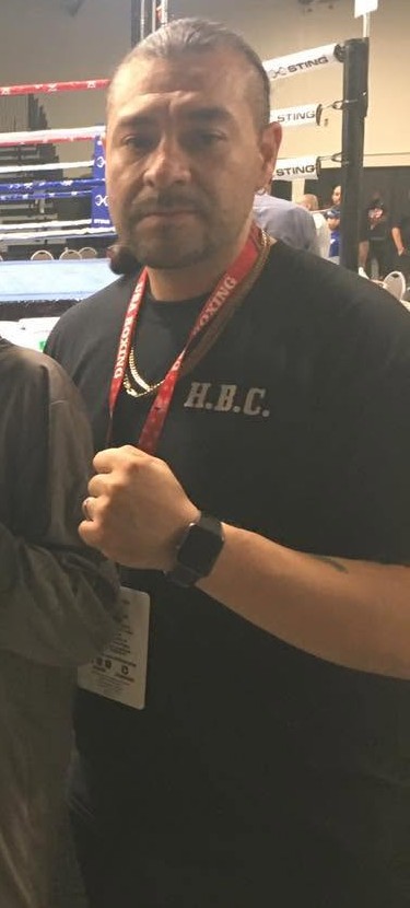 Coach Jesus Dominguez in an H.B.C. shirt and USA Boxing lanyard, fist raised at an indoor tournament.