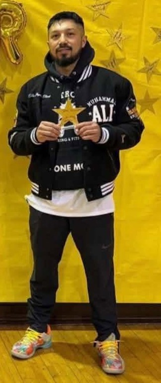 Coach Michael Jadd in a Muhammad Ali Boxing & Fitness varsity jacket holding a small star trophy in a school gym.