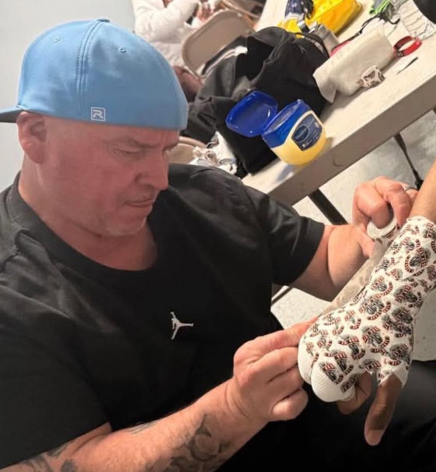 Coach Rafael Ruiz in a light-blue cap wrapping a boxer's hand backstage.