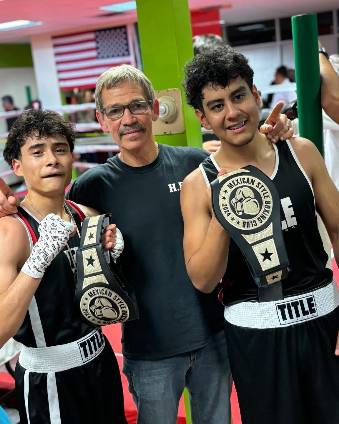 An older HBC coach standing between two teenage boxers holding 'Mexican Style Boxing Club 2024' championship belts.