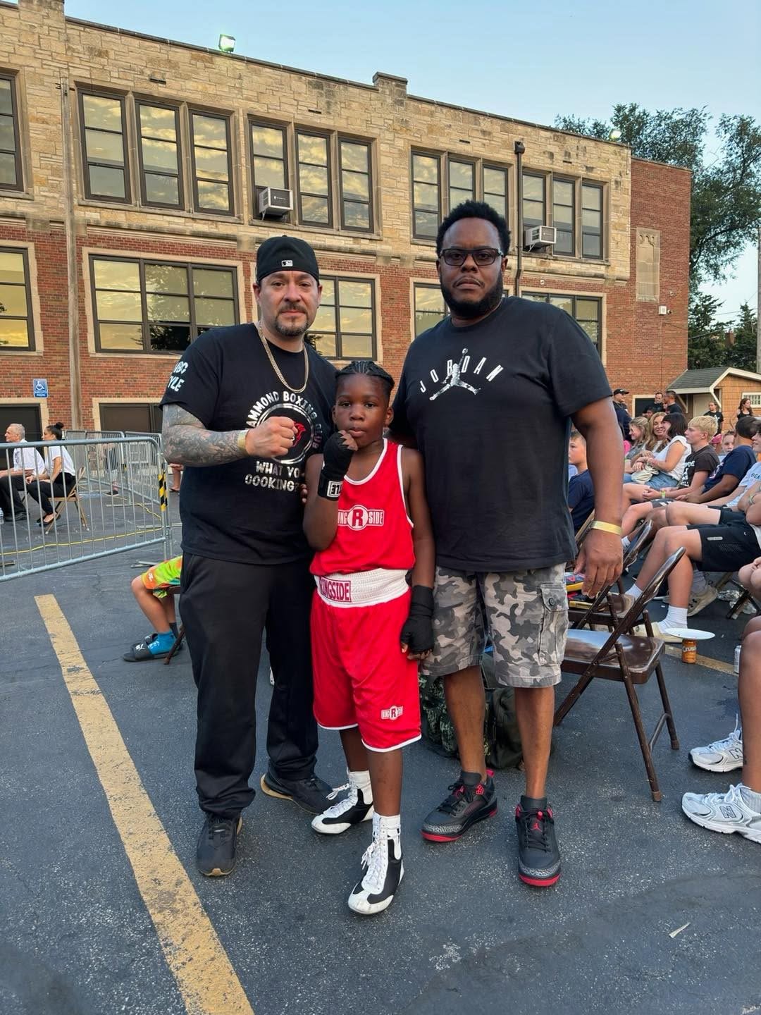 Two HBC coaches flanking a small boy boxer in a red Ringside uniform at an outdoor parking-lot event.