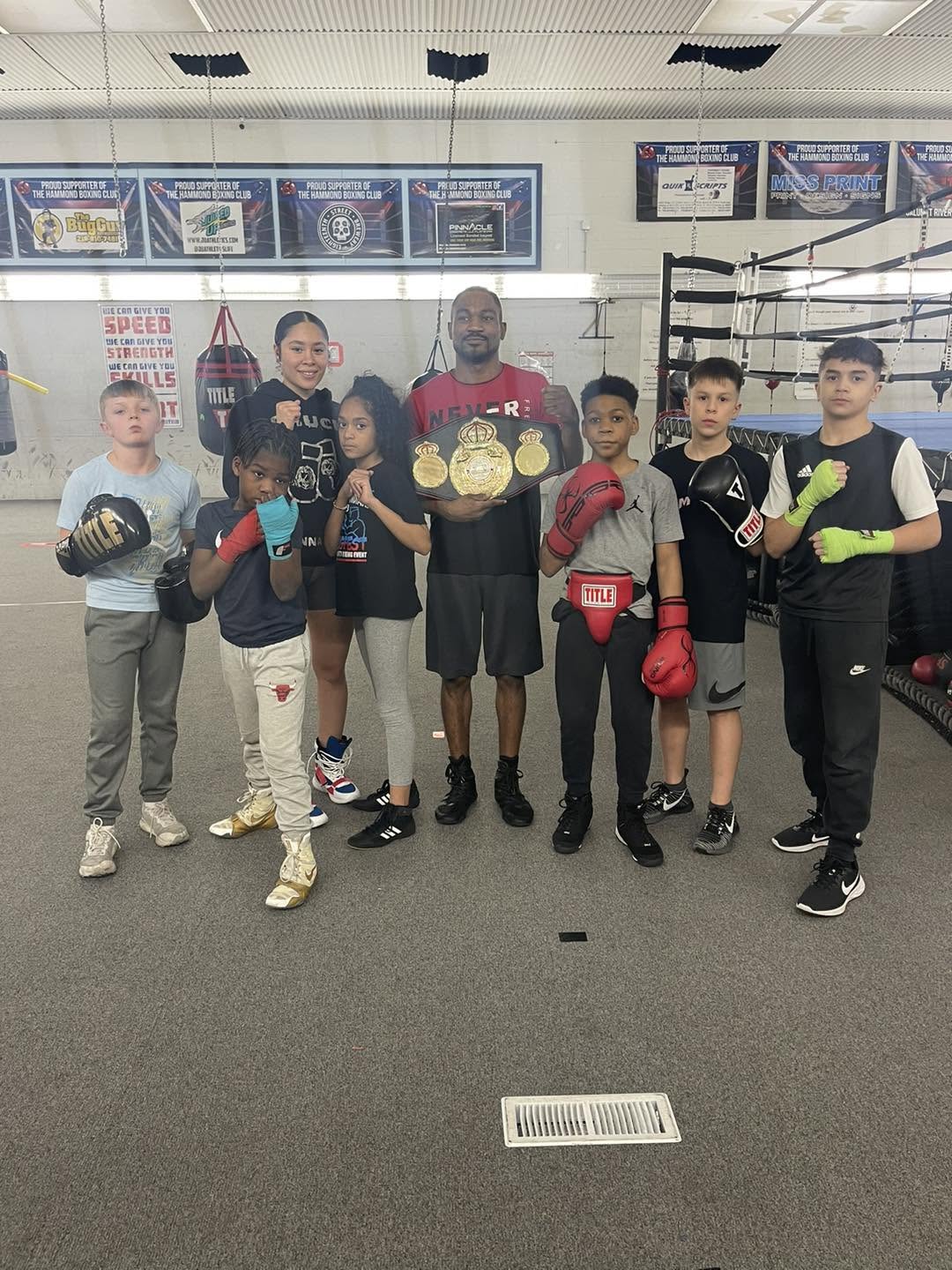 Two coaches with six youth boxers in gloves posed in the gym, one coach holding a championship belt, sponsor banners on the wall.