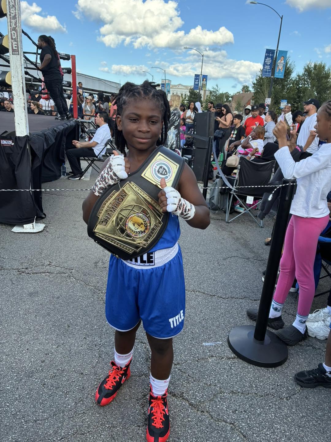 A young boy boxer in a blue TITLE uniform holding a 'Champion' belt at an outdoor street boxing event.