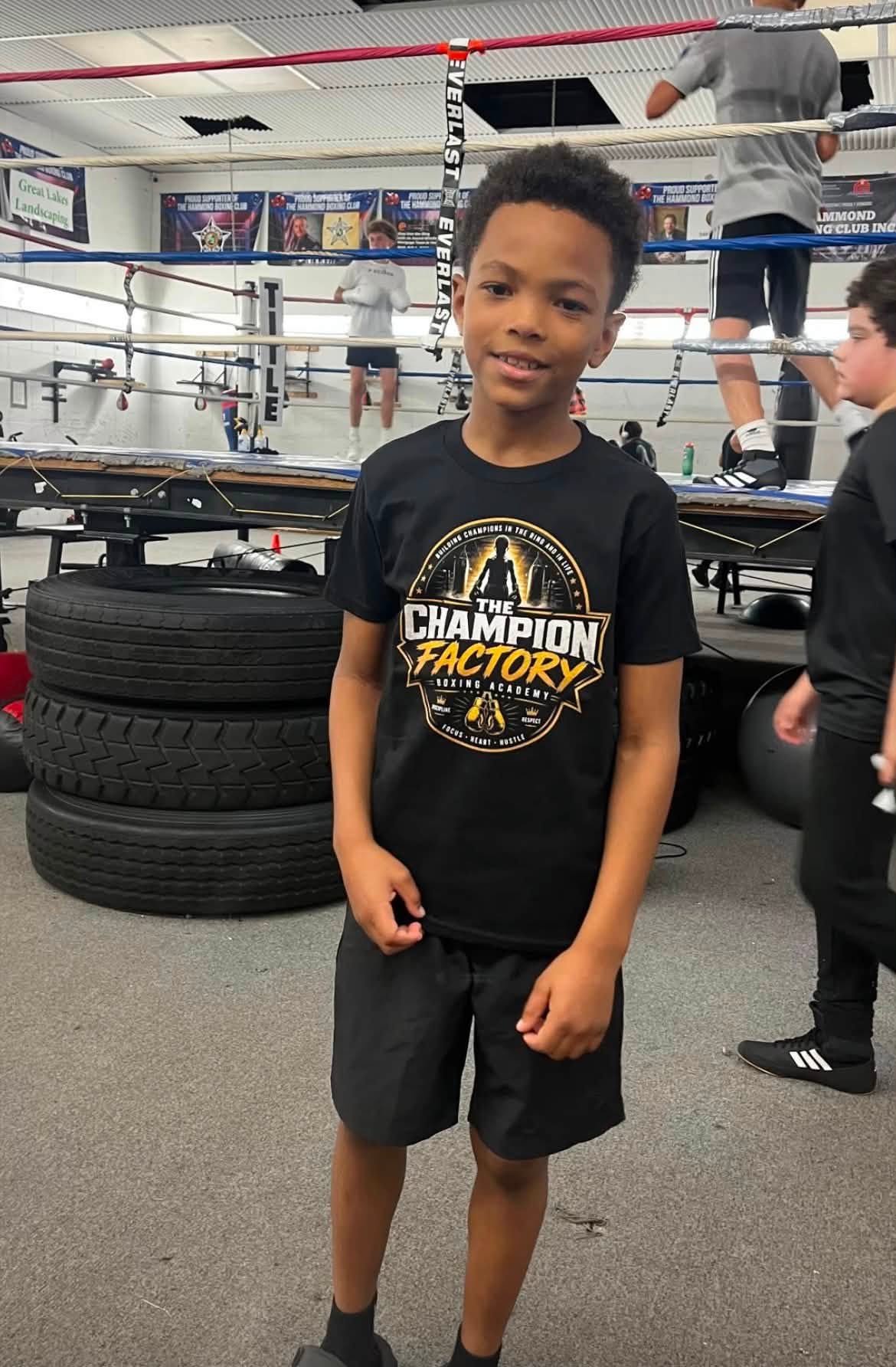 A young boy in a 'Champion Factory Boxing Academy' tee standing in the gym near the ring and training tires.