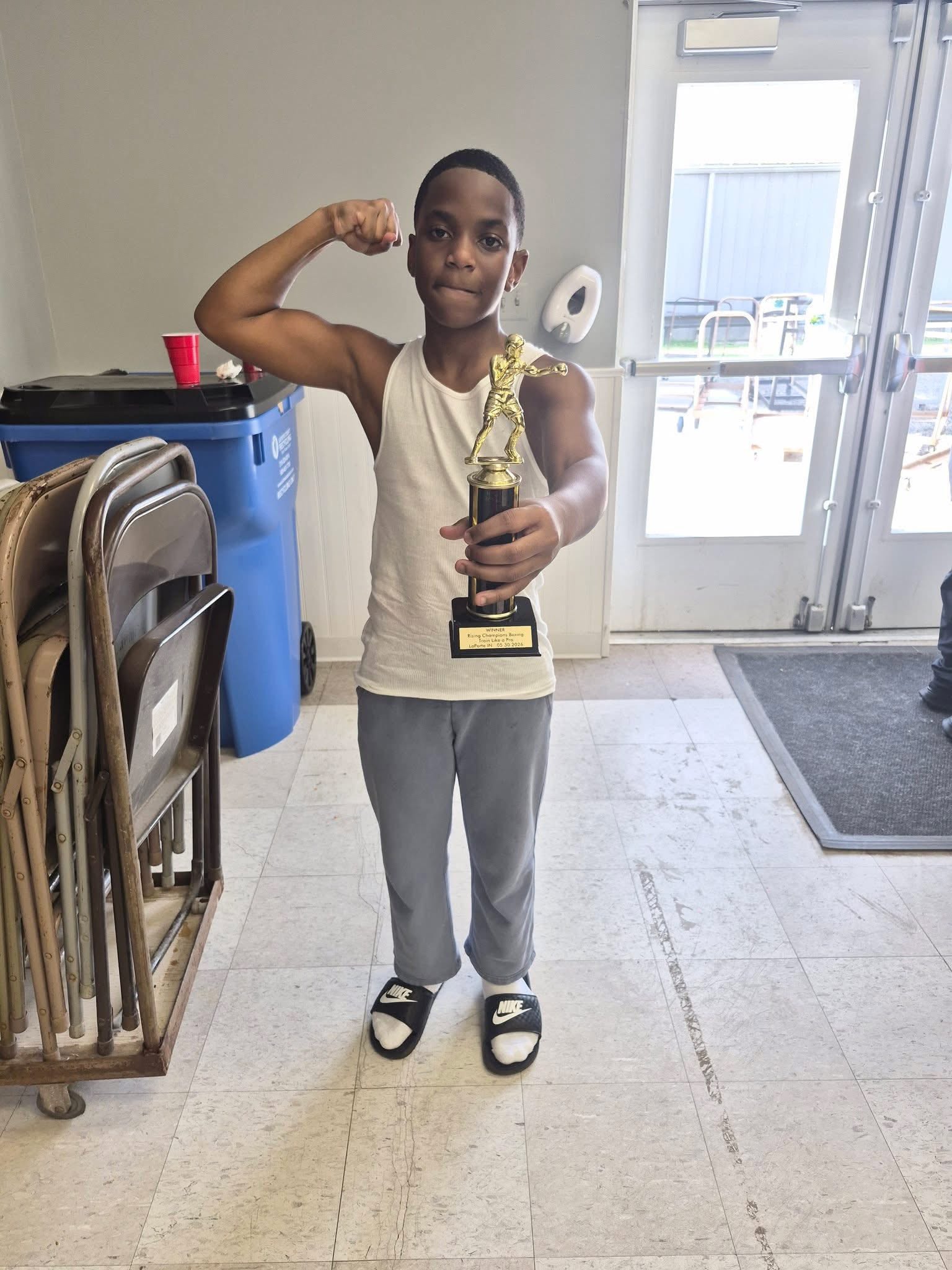 A young boy flexing while holding a small boxing trophy in a community-center lobby.