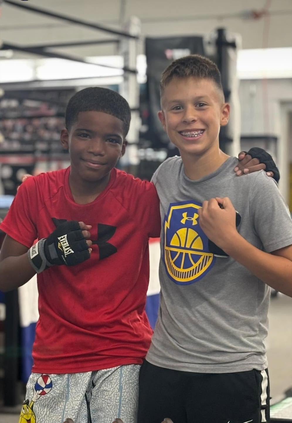 Two young boys in hand-wrap gloves, arms around each other, in front of the boxing ring.