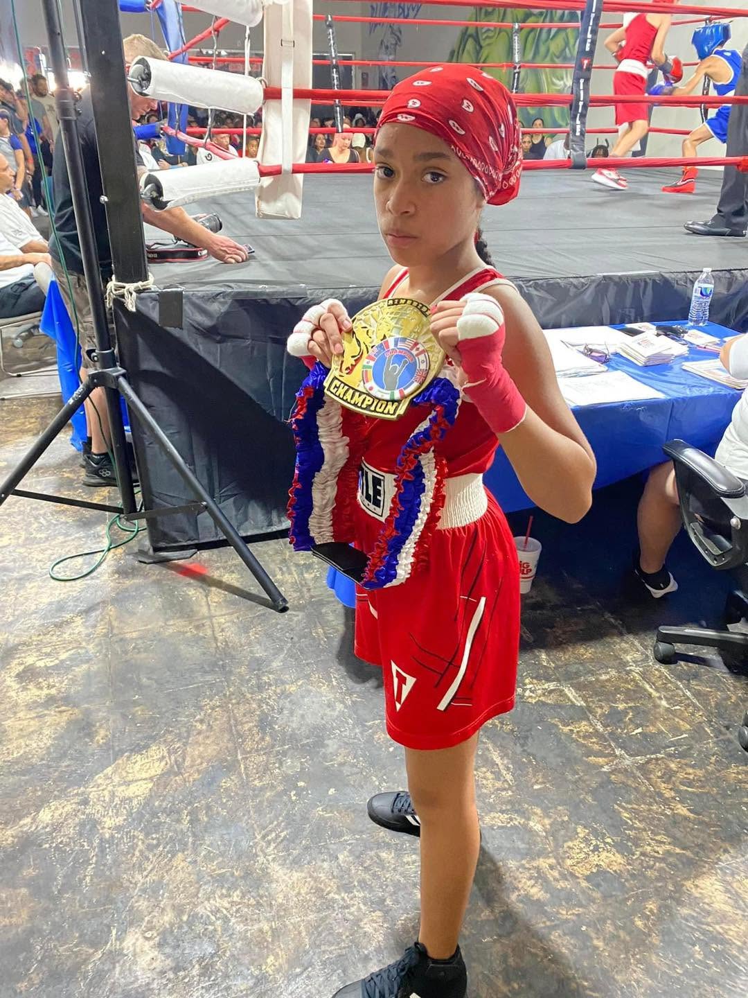 A young girl boxer in a red uniform and bandana holding a gold 'Champion' belt at a tournament.