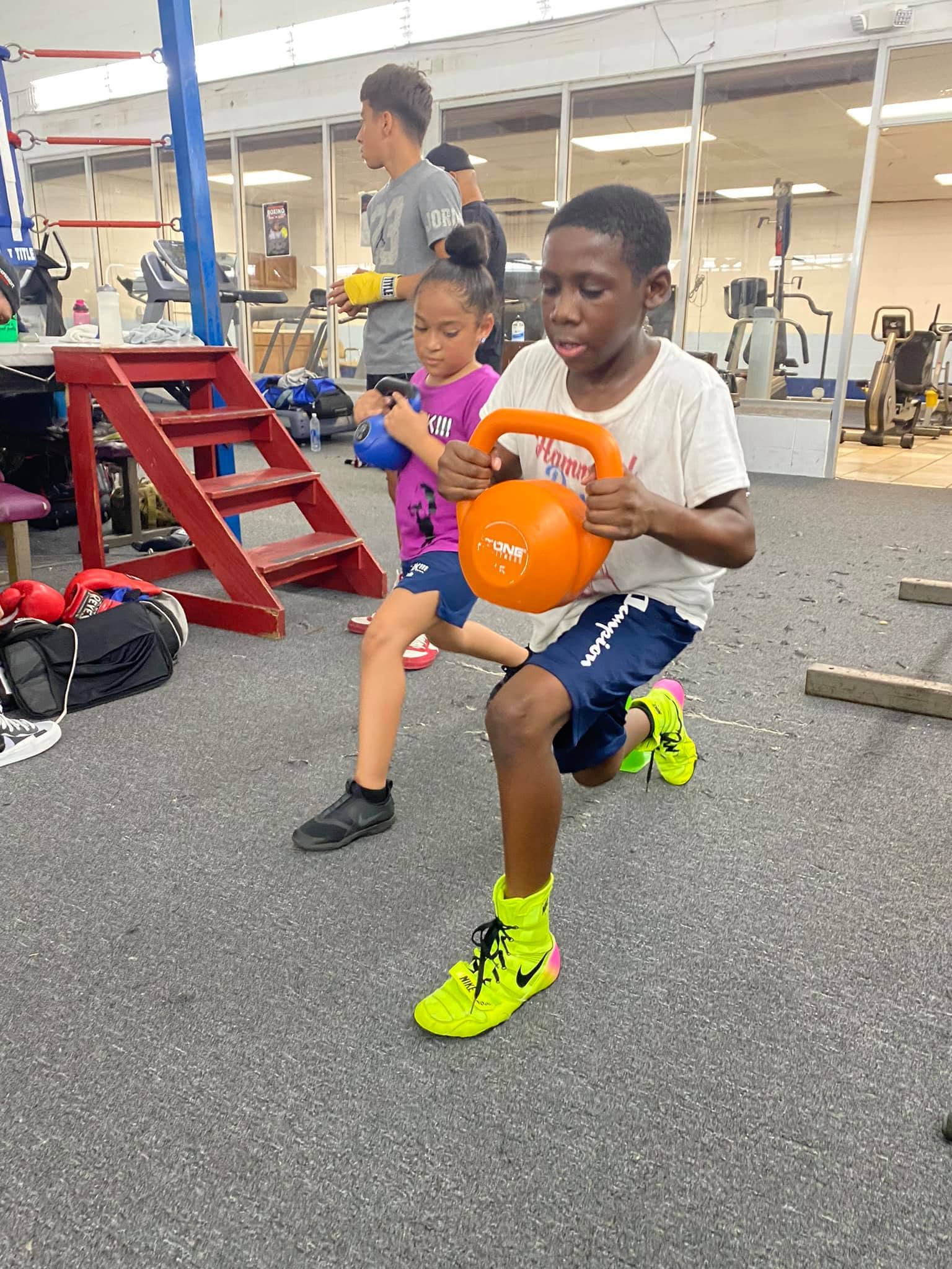 A young boy doing a lunge with an orange kettlebell during a youth workout, a young girl training behind him.