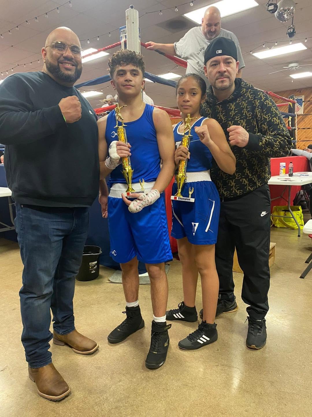 Two coaches with a teen boy and teen girl in blue uniforms, each holding a trophy, at an indoor tournament.